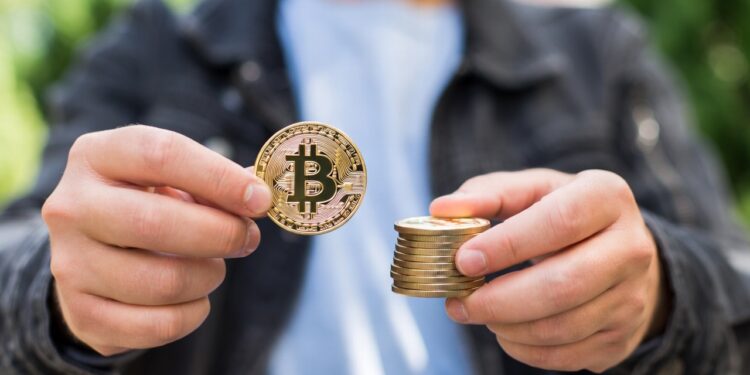 Hombre sostiene una moneda física de bitcoin en una mano y una pila de monedas también de bitcoin en la otra.