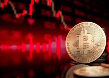 Moneda física de bitcoin dorada con gráficas rojas en el fondo.