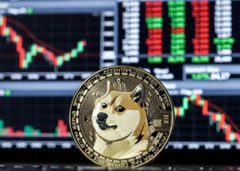 Moneda física de dogecoin en primer plano, al fondo un panel electrónico típico de Wall Street lleno de gráficos de velas rojas y verdes.