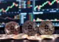 Varias monedas físicas de Ethereum en el fondo una pantalla con gráficos de velas bursátiles en verde y rojo.
