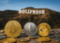 Varias monedas físicas de criptomonedas —Bitcoin, Ethereum y Dogecoin en primer plano— colocadas sobre una superficie, mientras al fondo se ve borroso el famoso cartel de “HOLLYWOOD” en una colina bajo un cielo despejado.