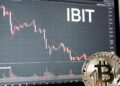 Una moneda física de bitcoin dorada y brillante apoyada sobre una superficie oscura; al fondo, una pantalla de trading con un gráfico y destaca en grande y en blanco el ticker «IBIT».