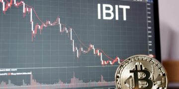 Una moneda física de bitcoin dorada y brillante apoyada sobre una superficie oscura; al fondo, una pantalla de trading con un gráfico y destaca en grande y en blanco el ticker «IBIT».