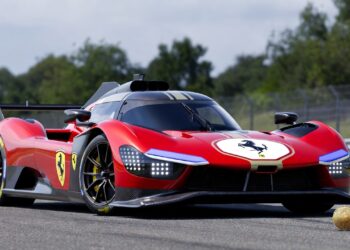 Un hiperrealista Ferrari 499P 2025 rojo con detalles aerodinámicos, alerón trasero y emblema del cavallino rampante, estacionado en una pista de carreras asfaltada con vallas y vegetación al fondo.