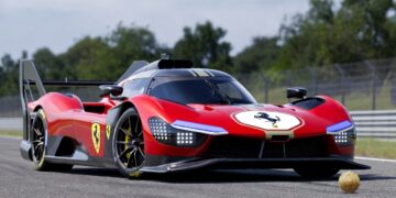 Un hiperrealista Ferrari 499P 2025 rojo con detalles aerodinámicos, alerón trasero y emblema del cavallino rampante, estacionado en una pista de carreras asfaltada con vallas y vegetación al fondo.