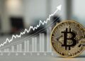 Una moneda física de bitcoin en primer plano a la derecha, con un gráfico de barras y una línea ascendente en el fondo que representa la inflación en EE. UU.