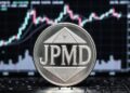 Una moneda metálica plateada con las letras JPMD en relieve en su centro con un fondo de una pantalla con gráficos de velas bursátiles.