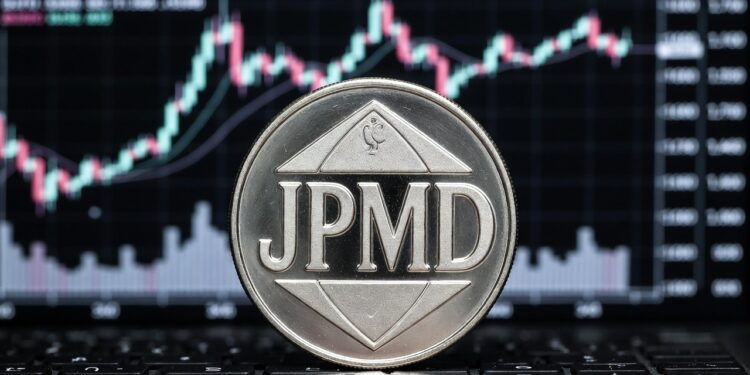 Una moneda metálica plateada con las letras JPMD en relieve en su centro con un fondo de una pantalla con gráficos de velas bursátiles.
