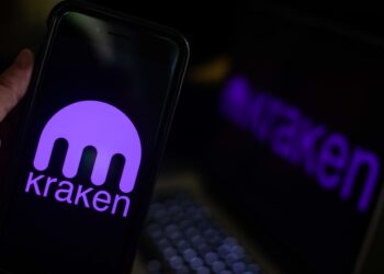 Una mano sostiene un smartphone en primer plano que muestra el logotipo de Kraken.