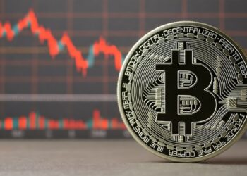 Una moneda física de bitcoin de color plateado con el logo de la B mayúscula en el centro. Detrás de la moneda, desenfocado, se observa un gráfico de velas rojas.