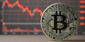 Una moneda física de bitcoin de color plateado con el logo de la B mayúscula en el centro. Detrás de la moneda, desenfocado, se observa un gráfico de velas rojas.