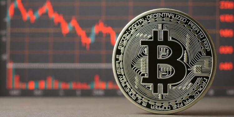 Una moneda física de bitcoin de color plateado con el logo de la B mayúscula en el centro. Detrás de la moneda, desenfocado, se observa un gráfico de velas rojas.