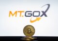 Moneda física de bitcoin dorada sobre un escritorio con el logo de Mt.Gox de fondo.