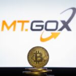 Moneda física de bitcoin dorada sobre un escritorio con el logo de Mt.Gox de fondo.
