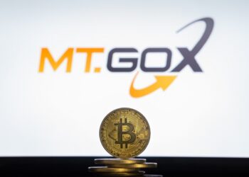 Moneda física de bitcoin dorada sobre un escritorio con el logo de Mt.Gox de fondo.