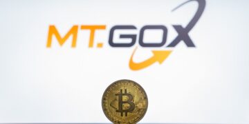 Moneda física de bitcoin dorada sobre un escritorio con el logo de Mt.Gox de fondo.