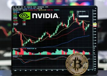 una moneda física de bitcoin dorada en primer plano sobre el teclado de una laptop. En la pantalla se muestra un gráfico bursátil con el logo oficial de NVIDIA.