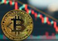 Bitcoin físico dorado en primer plano, ligeramente inclinado hacia la derecha, con un fondo desenfocado de un gráfico financiero de velas japonesas con velas rojas y verdes sobre un fondo oscuro, que parece indicar una tendencia a la baja.