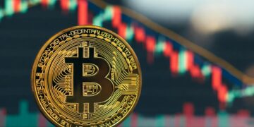 Bitcoin físico dorado en primer plano, ligeramente inclinado hacia la derecha, con un fondo desenfocado de un gráfico financiero de velas japonesas con velas rojas y verdes sobre un fondo oscuro, que parece indicar una tendencia a la baja.