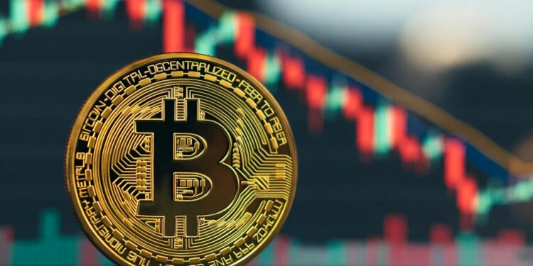 Bitcoin físico dorado en primer plano, ligeramente inclinado hacia la derecha, con un fondo desenfocado de un gráfico financiero de velas japonesas con velas rojas y verdes sobre un fondo oscuro, que parece indicar una tendencia a la baja.