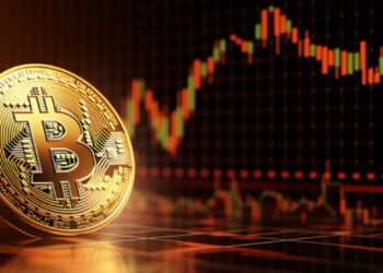 Moneda física de bitcoin dorada en un escritorio con gráficas de mercado en el fondo.