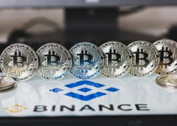 Una fila de monedas plateadas de Bitcoin alineadas horizontalmente sobre una superficie blanca con el logotipo azul de Binance y el texto "BINANCE" debajo.