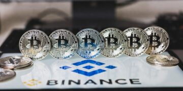 Una fila de monedas plateadas de Bitcoin alineadas horizontalmente sobre una superficie blanca con el logotipo azul de Binance y el texto "BINANCE" debajo.