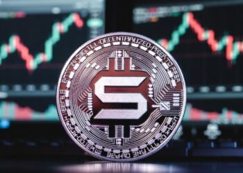 Moneda física de solana en primer plano apoyada sobre una superficie oscura, con un fondo de pantallas de trading bursátil.