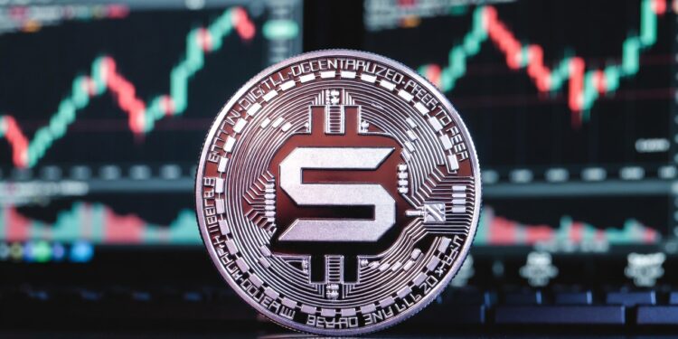 Moneda física de solana en primer plano apoyada sobre una superficie oscura, con un fondo de pantallas de trading bursátil.