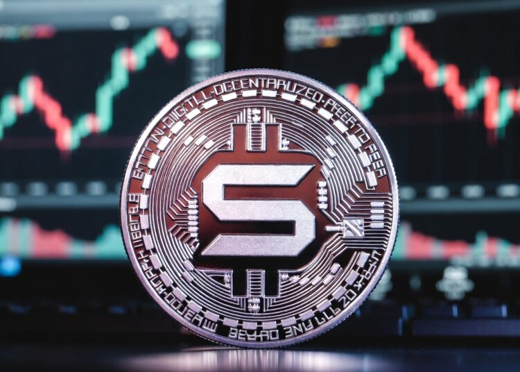 Moneda física de solana en primer plano apoyada sobre una superficie oscura, con un fondo de pantallas de trading bursátil.