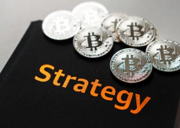 Varias monedas físicas de bitcoin plateadas sobre una carpeta negra con el nombre 'Strategy' en letras color naranja.