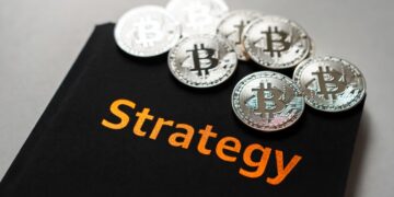 Varias monedas físicas de bitcoin plateadas sobre una carpeta negra con el nombre 'Strategy' en letras color naranja.