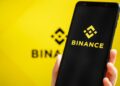 Una mano de tez blanca sujetando un teléfono inteligente que muestra el logo de Binance, y en el fondo, el logo de Binance difuminado.