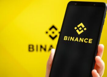 Una mano de tez blanca sujetando un teléfono inteligente que muestra el logo de Binance, y en el fondo, el logo de Binance difuminado.