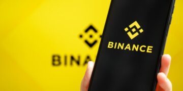 Una mano de tez blanca sujetando un teléfono inteligente que muestra el logo de Binance, y en el fondo, el logo de Binance difuminado.