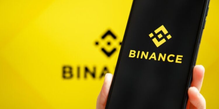 Una mano de tez blanca sujetando un teléfono inteligente que muestra el logo de Binance, y en el fondo, el logo de Binance difuminado.