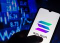 Una mano sujetando un teléfono inteligente que muestra el logo de Solana, y de fondo una pared de gráficas en color azul.