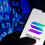 Una mano sujetando un telรฉfono inteligente que muestra el logo de Solana, y de fondo una pared de grรกficas en color azul.