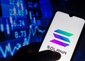 Una mano sujetando un teléfono inteligente que muestra el logo de Solana, y de fondo una pared de gráficas en color azul.