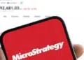 Un mรณvil con el logo de Microstrategy y en el fondo el precio de bitcoin.