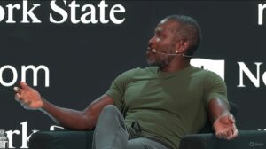 Arthur Hayes vistiendo una camiseta verde y pantalón gris, mientras habla en un escenario durante una conferencia.