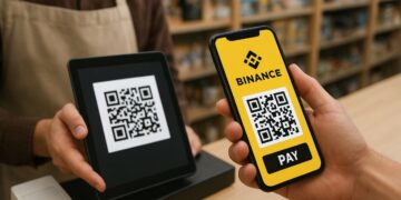 Una persona se encuentra en una tienda realizando un pago con su teléfono móvil. En la pantalla del celular se ve claramente el logotipo amarillo de Binance y un código QR con el botón “PAY” debajo. Frente a ella, un empleado sostiene una tableta negra que muestra otro código QR para recibir el pago. Ambos están en un mostrador de madera, con estantes llenos de productos desenfocados al fondo. La iluminación es cálida y natural, dando un ambiente acogedor y cotidiano, típico de una tienda moderna.