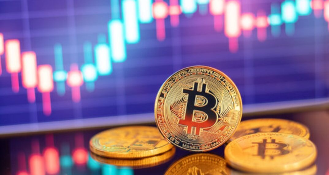 Monedas físicas de bitcoin doradas en un escritorio con gráficas de mercado en el fondo.