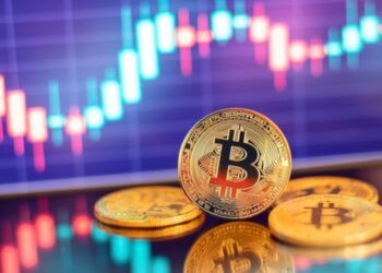 Monedas físicas de bitcoin doradas en un escritorio con gráficas de mercado en el fondo.