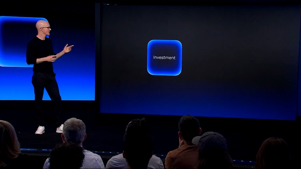 Brian Armstrong stands on a blue-lit stage, gesturing with his hands as he gives a presentation.