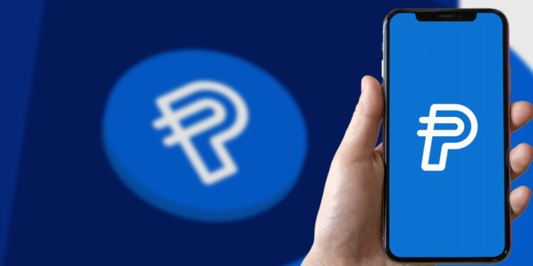Mano sosteniendo un teléfono inteligente que muestra el logo de PYUSD y en el fondo la moneda PYUSD.