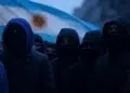 Encapuchados frente a una bandera de Argentina.