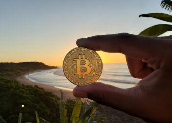 Una mano humana sosteniendo una moneda de bitcoin entre su dedo pulgar e índice y de fondo mar, sol y montañas de El Salvador.