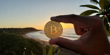 Una mano humana sosteniendo una moneda de bitcoin entre su dedo pulgar e índice y de fondo mar, sol y montañas de El Salvador.