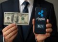 Empresario sostiene un billete de 100 dólares mientras muestra un teléfono móvil con el logo de Ripple y un botón de “comprar”.